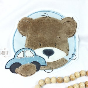 Könnte beinhalten: Weißer Body mit einem braunen Teddybär-Applikation und einem blauen Auto. Der Bär ist von einem hellblauen Kreis mit einer genähten Bordüre umgeben. Das Auto ist hellblau mit weißen Punkten.