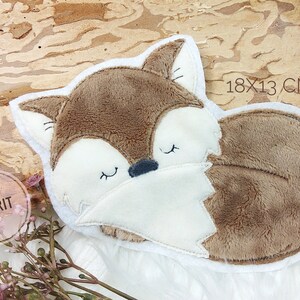 Könnte beinhalten: Ein fuchsformiges Kissen aus Plüsch in Braun und Weiß, 18 x 13 cm groß. Der Fuchs liegt zusammengerollt mit geschlossenen Augen auf einem Holzgrund. Das Kissen hat eine weiche, flauschige Textur.