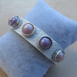 Puede incluir: Un brazalete de cuero blanco con cinco botones redondos, morados y plateados.