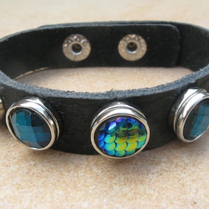 Può includere: Braccialetto in pelle nera con cinque accenti in metallo argentato, ognuno con una gemma blu e verde iridescente diversa.