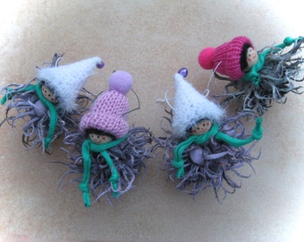 May include: Four whimsical handmade ornaments. Each features a smiling face, a knitted hat in white or pink, and a green scarf. The figures are attached to a cluster of gray and purple decorative elements, perfect for holiday decor.