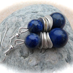 Könnte beinhalten: Ohrringe mit tiefblauen, kugelförmigen Perlen, kombiniert mit silberfarbenen, blütenblattförmigen Elementen. Die Ohrringe haben ein einfaches Haken-Design. Der Schmuck wird auf einer grauen Steinoberfläche präsentiert.