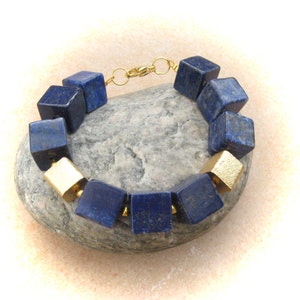 Può includere: Bracciale con perline a forma di cubo blu scuro e oro alternate. Il bracciale è collegato con una catena e una chiusura color oro. Le perline sono disposte in un motivo circolare.