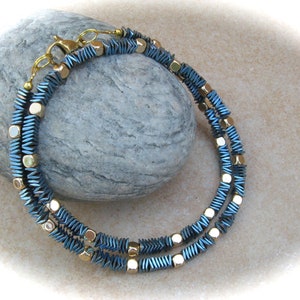 Può includere: Una collana a doppio filo realizzata con perline metalliche blu e dorate. Le perline sono sfaccettate e hanno una forma geometrica.
