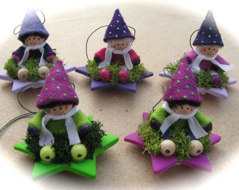 May include: Five whimsical star-shaped ornaments, each featuring a felt gnome figure. The gnomes wear colorful outfits, including purple hats with green polka dots, and sit on a bed of green moss. The stars are painted in shades of purple and green.
