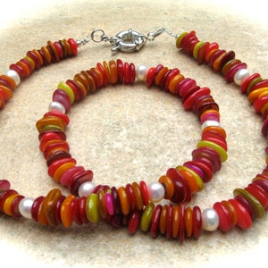 Op de afbeelding: Een meerstrengs ketting gemaakt van gelaagde, kleurrijke schelp kralen in tinten rood, oranje, geel en groen. De ketting is versierd met witte parels en een zilveren sluiting.