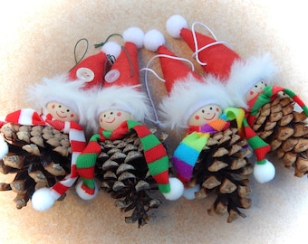May include: A collection of handmade Christmas ornaments. Each ornament features a pine cone body, a painted wooden head, and a festive scarf. They are adorned with red Santa hats trimmed with white faux fur and white pom-poms.