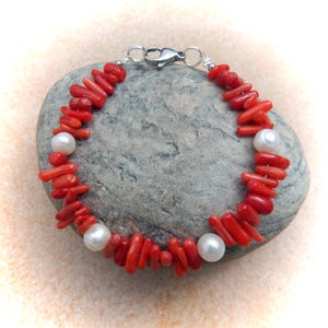 Può includere: Un bracciale caratterizzato da perle di corallo rosso irregolari a forma di ramo alternate a perle bianche lisce e rotonde. Il gioiello è fissato con una chiusura a moschettone in metallo argentato, creando un cerchio di colori e consistenze contrastanti.