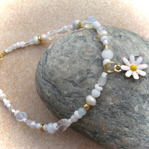 Pode incluir: Pulseira de contas brancas e douradas com um amuleto de margarida branca. A pulseira é feita com contas brancas, contas douradas e uma conta de cristal transparente. O amuleto de margarida tem um centro amarelo.