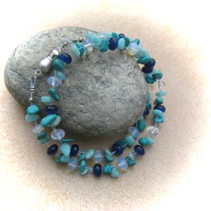 Op de afbeelding: Een meerstrengige ketting gemaakt met turquoise, blauwe en witte kralen. De ketting is gemaakt met verschillende vormen en maten kralen, waaronder ronde, ovale en onregelmatige vormen. De ketting is geregen op een zilveren ketting.