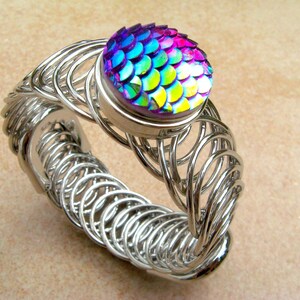 May include: A silver wire bracelet with a large iridescent button in the center. The button has a scale-like pattern and is a mix of purple, blue, green, and yellow.