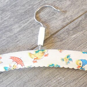 May include: A vintage clothing hanger with a white handle and a metal hook. The hanger is covered in a fabric with a scalloped edge, featuring a repeating pattern of cartoon ducks, umbrellas, and flowers in pastel colors.