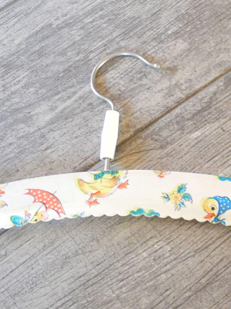 May include: A vintage clothing hanger with a white handle and a metal hook. The hanger is covered in a fabric with a scalloped edge, featuring a repeating pattern of cartoon ducks, umbrellas, and flowers in pastel colors.