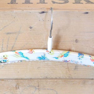 May include: A vintage white wooden clothes hanger with a decorative floral and bird pattern in blue, yellow, and red. The hanger has a metal hook and a scalloped edge.