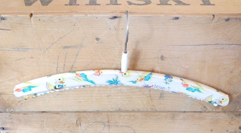 May include: A vintage white wooden clothes hanger with a decorative floral and bird pattern in blue, yellow, and red. The hanger has a metal hook and a scalloped edge.