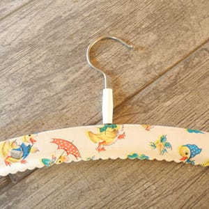 May include: A vintage-style clothing hanger with a white base and a scalloped edge. The hanger is decorated with a repeating pattern of cartoon ducks, umbrellas, and floral elements in shades of yellow, blue, and red. The hook is silver-colored.