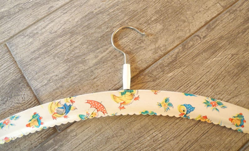 May include: A vintage-style clothing hanger with a white base and a scalloped edge. The hanger is decorated with a repeating pattern of cartoon ducks, umbrellas, and floral elements in shades of yellow, blue, and red. The hook is silver-colored.