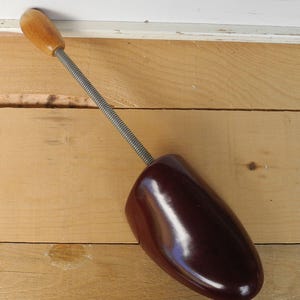 May include: A vintage shoe stretcher. The shoe form is a dark brown, glossy material. It has a silver-coloured spring and a wooden handle. The shoe stretcher is on a light brown wooden surface.