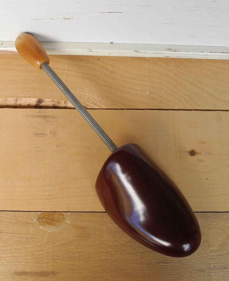 May include: A vintage shoe stretcher. The shoe form is a dark brown, glossy material. It has a silver-coloured spring and a wooden handle. The shoe stretcher is on a light brown wooden surface.