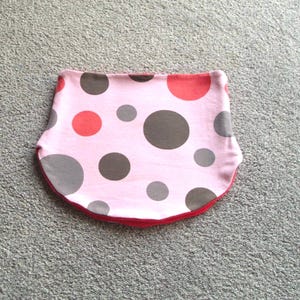 Könnte beinhalten: Ein rosa Lätzchen mit einem Muster aus großen grauen, roten und braunen Punkten. Das Lätzchen hat eine rote Einfassung und ist aus weichem Stoff. Das Lätzchen soll Spritzer auffangen und die Kleidung schützen.