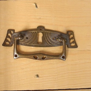 Könnte beinhalten: Antiker Schubladengriff aus Messing mit Schlüsselloch und dekorativem floralem Design. Der Griff ist an einer hellen Holzoberfläche befestigt. Der Schubladengriff ist im Vintage-Stil gehalten und hat ein angelaufenes Finish.