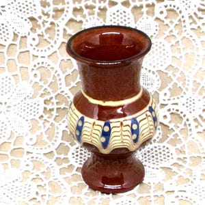 Könnte beinhalten: Kleine, braune Keramikvase mit ausgestelltem Oberteil und rundem Boden. Die Vase hat ein dekoratives Band mit cremefarbenen und blauen Akzenten in der Mitte. Die Vase ist etwa 13 cm hoch und hat eine glänzende Oberfläche.