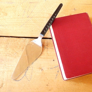 Könnte beinhalten: Ein Kuchenheber aus Edelstahl mit schwarzem Griff liegt auf einer Holzoberfläche neben einem roten Buch. Der Heber hat eine breite, dreieckige Klinge und einen Griff mit kleinen, kreisförmigen Vertiefungen.