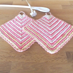 Könnte beinhalten: Zwei handgefertigte, quadratische Topflappen aus Häkelarbeit mit einem rosa, weißen und gelben Streifenmuster. Jeder Topflappen hat eine kleine Schlaufe zum Aufhängen. Ein weißer Holzlöffel und eine kleine weiße Löffelablage liegen auf der Holzoberfläche.
