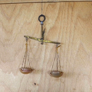 Könnte beinhalten: Antike Messingwaage mit zwei hängenden Schalen und einem zentralen Arm. Die Waage ist an einem Ring aufgehängt und vor einem Holzgrund befestigt. Die Schalen sind hellbraun.