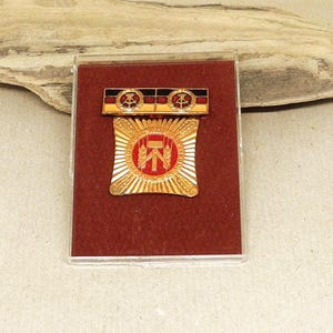 Könnte beinhalten: Eine goldfarbene Vintage-Medaille mit einem rot-weißen Emblem, in einem transparenten Kunststoffetui. Die Medaille hat ein Sonnenstrahlendesign mit einem zentralen Emblem und einer kleineren Leiste darüber mit zwei Symbolen. Der Hintergrund ist tiefrot.