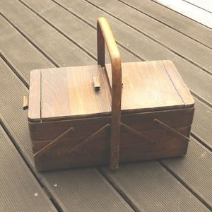 Könnte beinhalten: Ein rechteckiger Nähkasten aus dunkelbraunem Holz mit Klappdeckel und einem zentralen vertikalen Griff. Die Box verfügt über einen ausziehbaren Seitenmechanismus im Akkordeon-Stil mit diagonalen Holzleisten und einem kleinen Holzknopf.