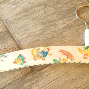May include: A vintage clothing hanger with a metal hook and a white handle. The hanger is covered in a cream-colored fabric with a scalloped edge and a repeating pattern of ducks, umbrellas, and flowers.