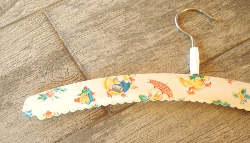 May include: A vintage clothing hanger with a metal hook and a white handle. The hanger is covered in a cream-colored fabric with a scalloped edge and a repeating pattern of ducks, umbrellas, and flowers.
