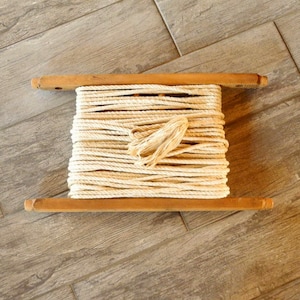 Könnte beinhalten: Eine Holzspule gefüllt mit beigem Seil. Die Spule hat an jedem Ende zwei Holzstäbe. Das Seil ist fest um die Mitte der Spule gewickelt. Die Spule liegt auf einer Holzoberfläche.