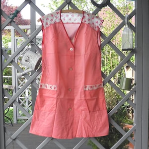 Könnte beinhalten: Eine vintage korallenfarbene Schürze mit einem weißen und floral gemusterten Joch und Taschenbesatz. Die Schürze hat einen V-Ausschnitt, eine Knopfleiste vorne und zwei Vordertaschen. Sie hängt an einem Holzbügel an einem grauen Gitterzaun.