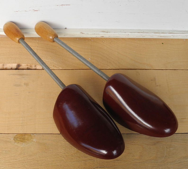 May include: A pair of vintage shoe trees. They have wooden handles and dark burgundy shoe forms. The metal springs connect the handles to the shoe forms. These are designed to maintain the shape of shoes.