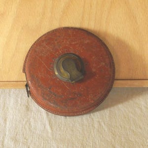 Könnte beinhalten: Ein rundes, vintage-artiges Maßband in einem abgenutzten, rotbraunen Ledergehäuse. In der Mitte befindet sich ein runder, messingfarbener Metallknopf zum Aufrollen. Auf der Lederoberfläche des Messwerkzeugs ist ein schwacher Prägetext zu sehen.