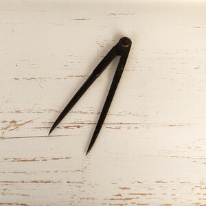 Könnte beinhalten: Ein dunkler, verwitterter Zirkel aus Metall mit zwei spitzen Schenkeln, die durch einen zentralen Drehpunkt verbunden sind. Das Werkzeug hat eine matte, gealterte Oberfläche mit sichtbaren Gebrauchsspuren und Oxidation, ein schlichtes Design.