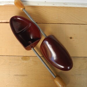 May include: Two vintage shoe trees, made of dark red plastic and wood, are displayed on a light brown wooden surface. The shoe trees have a spring mechanism and are designed to maintain the shape of shoes.
