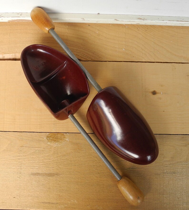 May include: Two vintage shoe trees, made of dark red plastic and wood, are displayed on a light brown wooden surface. The shoe trees have a spring mechanism and are designed to maintain the shape of shoes.