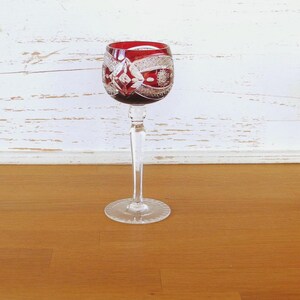 Könnte beinhalten: Ein rubinrotes Weinglas mit aufwendigen Schliffmustern. Das Glas hat einen langen, klaren Stiel und einen kreisförmigen Fuß. Die detaillierten Muster auf der Schale des Glases verleihen ihm eine elegante Note. Perfekt zum Servieren von Wein.