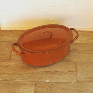 Könnte beinhalten: Eine ovale, rostfarbene Emaille-Auflaufform mit passendem Deckel. Die Form hat zwei seitliche Griffe und einen kleinen Griff am Deckel. Das Kochgeschirr steht auf einer Holzoberfläche.