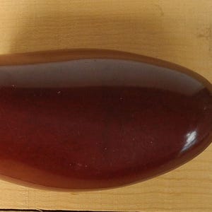 May include: A dark burgundy shoe tree with a glossy finish. The shoe form is oval-shaped and attached to a metal spring. The background is a light brown wooden surface.