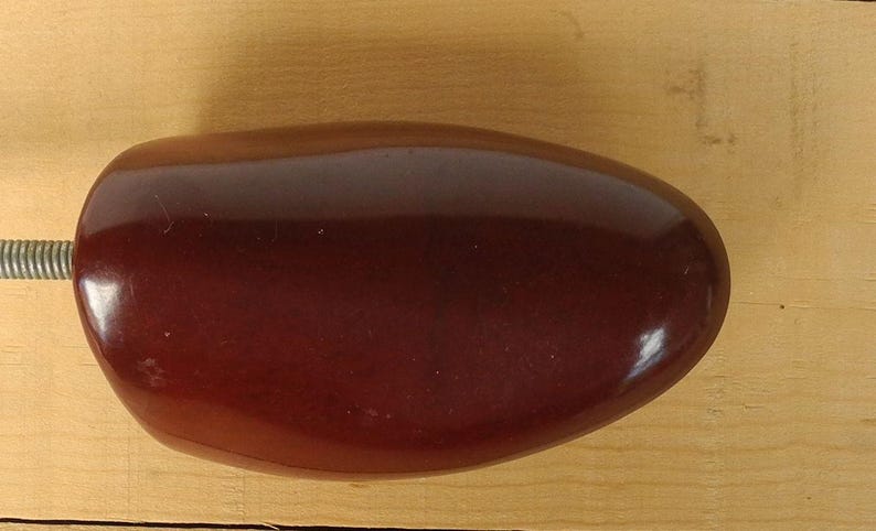 May include: A dark burgundy shoe tree with a glossy finish. The shoe form is oval-shaped and attached to a metal spring. The background is a light brown wooden surface.