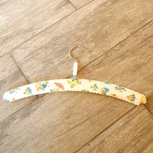 May include: A vintage, padded clothes hanger with a metal hook. The hanger is covered in a cream-colored fabric with a scalloped edge and a repeating pattern of cartoon ducks, umbrellas, and flowers.