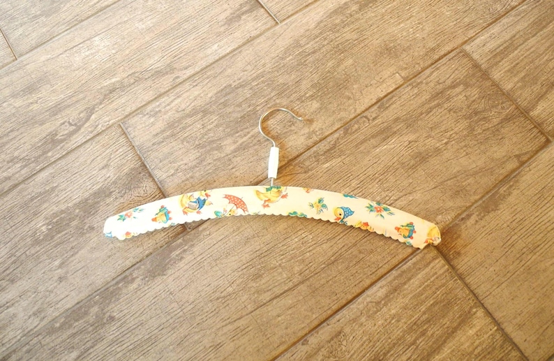 May include: A vintage, padded clothes hanger with a metal hook. The hanger is covered in a cream-colored fabric with a scalloped edge and a repeating pattern of cartoon ducks, umbrellas, and flowers.