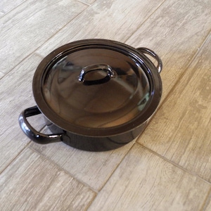 Könnte beinhalten: Ein schwarzer Emaille-Kochtopf mit passendem Deckel. Der Topf hat zwei seitliche Griffe und einen kleinen Griff am Deckel. Der Topf steht auf einer hellen, holzartig aussehenden Oberfläche. Dieses Kochgeschirr eignet sich für verschiedene Kochbedürfnisse.