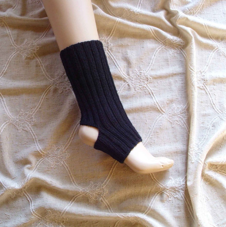 May include: Black knitted leg warmers with a cut-out at the heel. The leg warmers are shown on a mannequin leg against a beige fabric background.