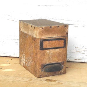 Könnte beinhalten: Eine Vintage-Holzkiste mit Scharnierdeckel und Metall-Etikettenhalter. Die Kiste hat eine abgenutzte Oberfläche, die Gebrauchsspuren aufweist. Ein Metallgriff befindet sich auf der Vorderseite. Die Kiste wird wahrscheinlich zur Aufbewahrung verwendet.