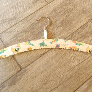 May include: A vintage clothing hanger with a cream-colored base and a scalloped edge. The hanger is decorated with a colorful pattern of birds, flowers, and other whimsical designs. The metal hook is silver.